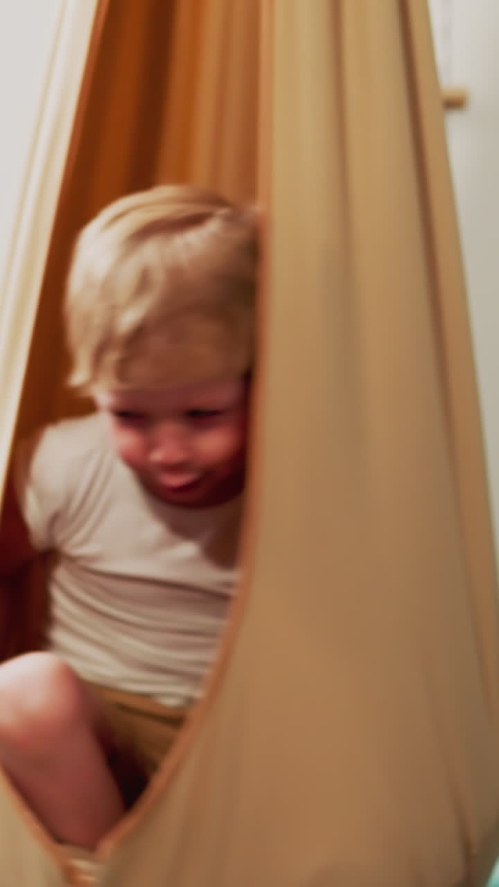 Happy little boy sits in turning hammock chair in home sports gym slow motion. Toddler kid swings in comfortable hanging textile cocoon in playing room