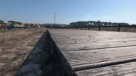 Tres-Personas-Caminan-A-Lo-Lejos-Sobre-Los-Tablones-De-Madera-Del-Paso-Elevado-Con-Barandillas-De-Madera-Viejas-Sobre-Suelo-Arenoso-En-Dirección-A-La-Ciudad,-Tomadas-Desde-Abajo-Girando-A-La-Derecha-Lentamente-Pontevedra,-Galicia,-España