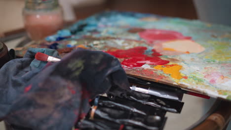 mesa con paleta de colores y pinceles. artista enjuaga pincel en vidrio lleno de agua quitando pintura. herramientas esenciales para la fabricación de arte