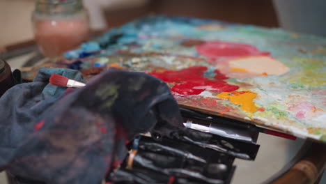 table with colours palette and brushes. artist rinses paintbrush in glass filled with water removing paint. essential tools for art manufacturing