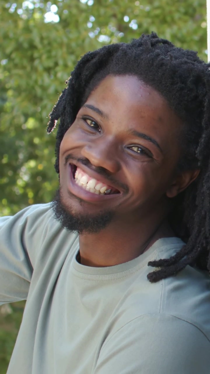 Vídeo vertical: Homem de camiseta verde fazendo contato visual, abrindo um sorriso perto de poste branco de varanda, com árvores ao fundo.