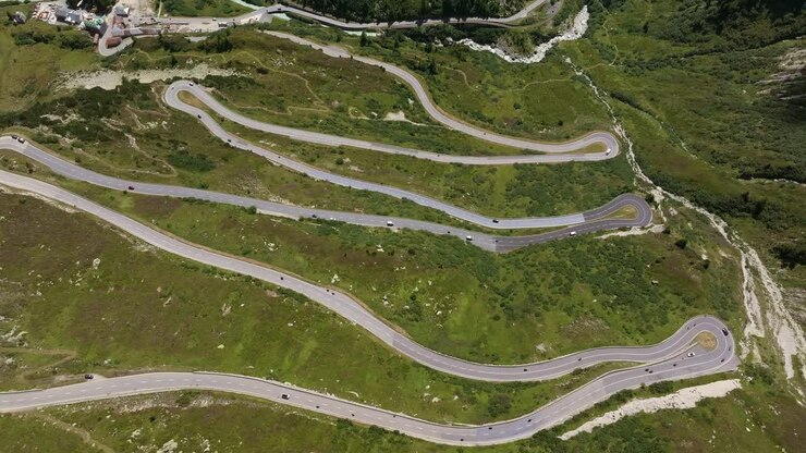 Ripresa aerea di un passo di montagna con numerosi tornanti stretti che si snodano attraverso un paesaggio alpino verde. Auto e camion percorrono la strada tortuosa.