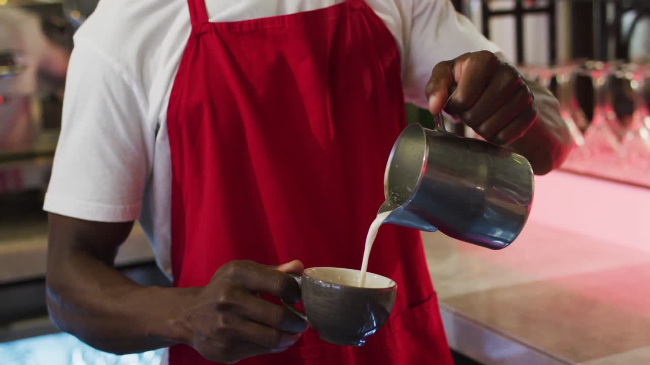sección media de un barista afroamericano vertiendo leche de un frasco en una taza de café en una cafetería