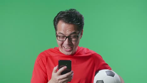 primer plano de un hombre asiático con una pelota gritando gol celebrando mientras aplaude el fútbol en el teléfono inteligente en el fondo de la pantalla verde