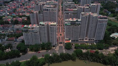 dramatic drone fly in over central road between tall buildings of a massive symmetrical housing estate on sunny day featuring river, towers, rooftops, gardens, swimming pool and tennis courts