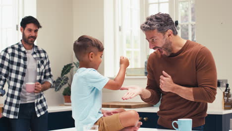 same sex family with two dads in kitchen with son sitting on counter playing rock paper scissors