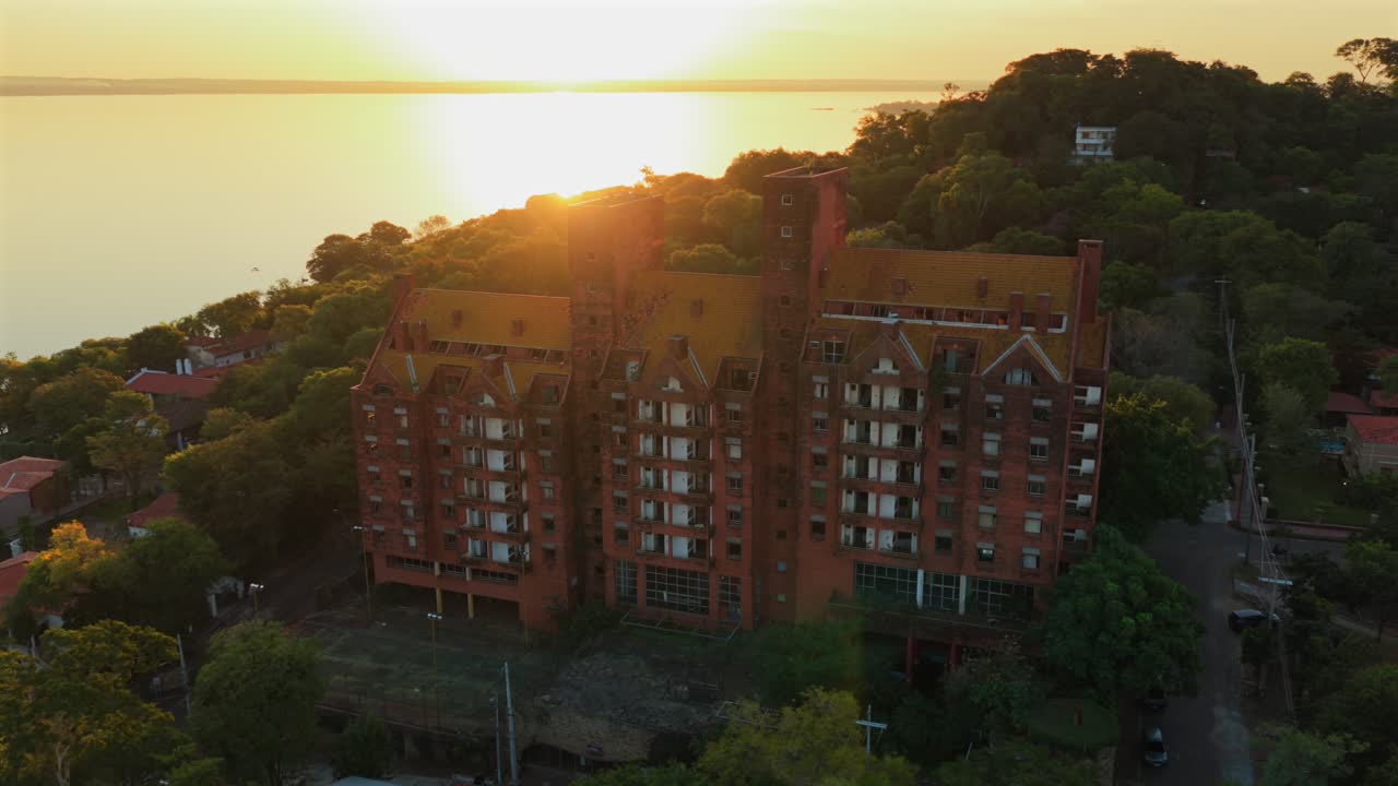 Drone video capturing stunning aerial view of San Bernardino, Paraguay, showcasing vibrant sunset hues, serene lakeside, and lush greenery surrounding a grand brick building