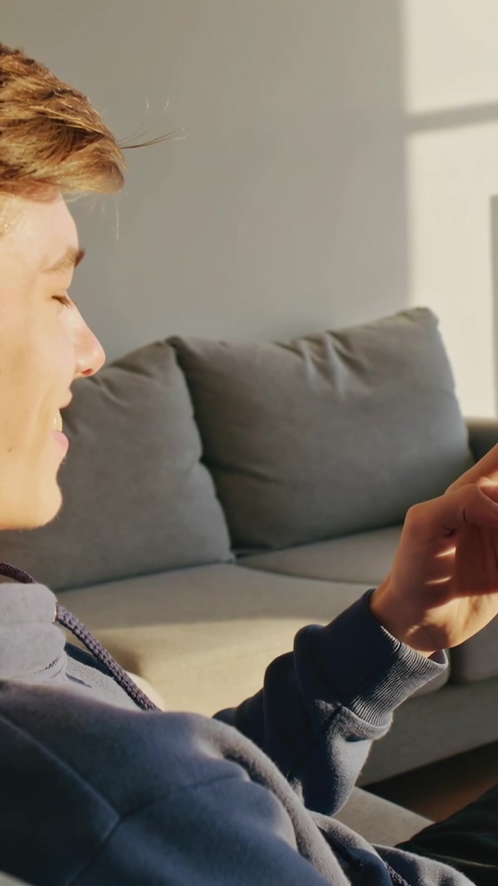 Vertikal video: Holder telefon, mandlig teenager i mørk hættetrøje trykker på skærm på sofa derhjemme, plads til tekst.