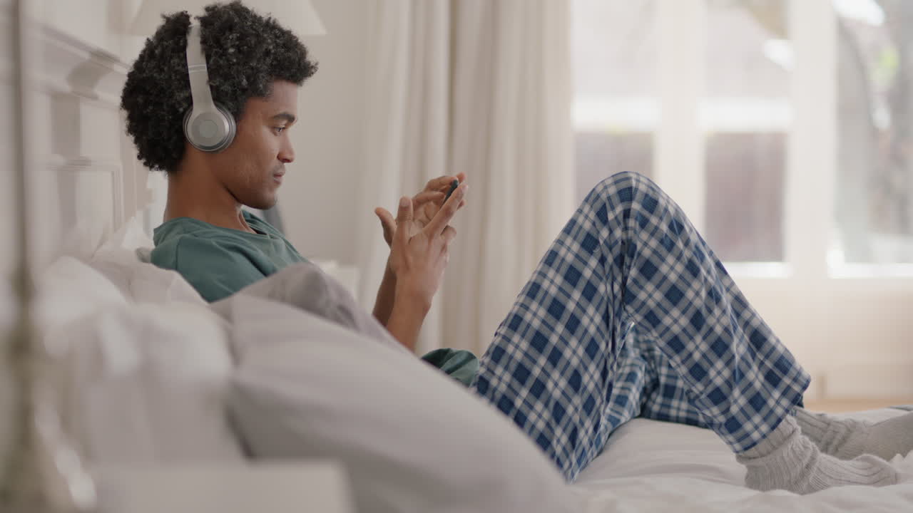 giovane uomo di razza mista che usa lo smartphone ascolta musica indossa le cuffie naviga online si rilassa a letto condivide lo stile di vita sui social media si gode la mattina del fine settimana