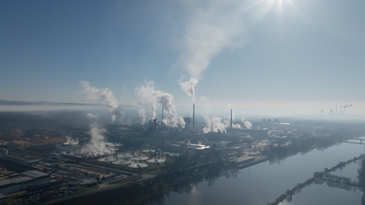 Vista aerea con drone che mette in risalto un'imponente ciminiera centrale e una densa colonna di vapore sopra il complesso industriale di Štětí, con capannoni di produzione e piazzali circostanti sottostanti