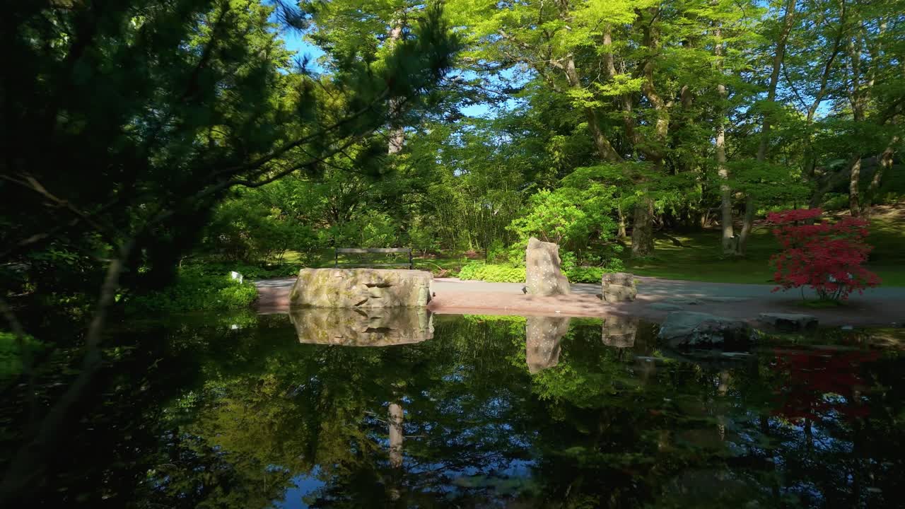 Movimento lento de câmera para frente revela uma ponte de pedra refletida em um lago tranquilo, cercada por árvores exuberantes e azaléias no Jardim Botânico de Gotemburgo.