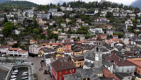 Aerial-flyover-from-over-the-rooftops-of-Ascona-in-Ticino,-Switzerland-towards-the-shores-of-Lago-Maggiore-with-a-boat-departing-the-city-waterfront-and-cruising-into-the-lake's-waters