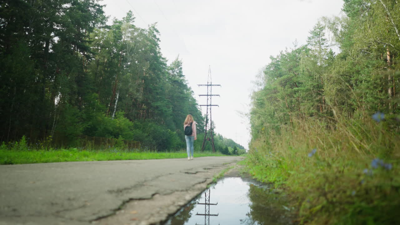 Woman walking in distance on quiet rural tarred road with visible potholes, surrounded by green forest and overcast sky, creating a serene yet rugged atmosphere with power lines stretching ahead