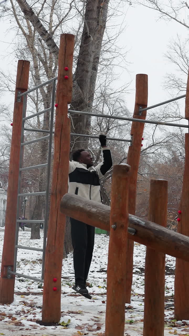 색 겨울 재을 입은 젊은이가 눈 인 공원에서 나무로 된 야외 운동 장비를 등반하며, 벌거벗은 나무와 추운 분위기는 겨울 훈련 동안의 결단력과 체력을 강조합니다.