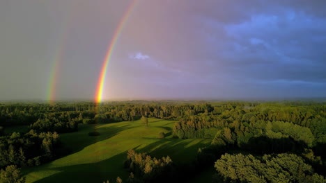 Cinematic-aerial-footage-of-a-double-rainbow-over-a-grassy-landscape-with-trees-on-a-cloudy-day,-Drone