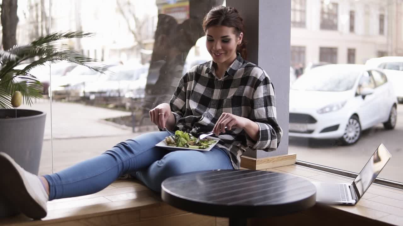 The girl sits leaning against the window. A plate on her legs. She mixes the green leaves of the salad with a fork and knife. Tastes the food and enjoys it. Slow motion