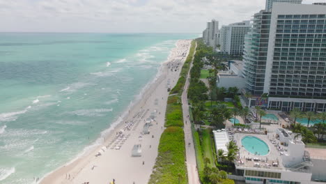 Forwards-fly-above-sea-coast.-People-enjoying-sunny-day-on-sand-beach.-Vacation-resort-with-palm-trees-and-luxurious-hotels.-Miami,-USA