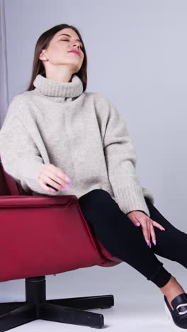 young beautiful long-haired brunette wearing warm sweater, tight pants and stylish shoes sitting in red armchair indoors. model posing in the studio. vertical video