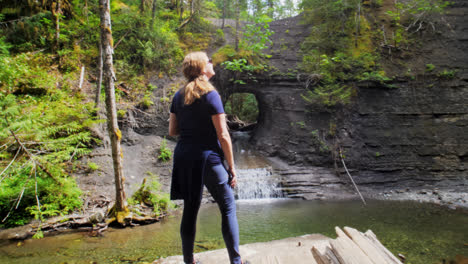 woman standing at lakeside looking at rock hole waterfall in forest, port alberni british columbia canada