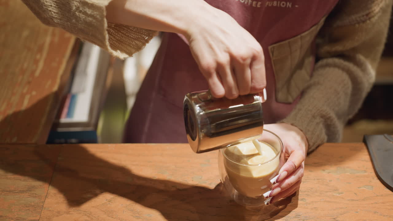 Close up of bartender in sweater and apron pouring milk into coffee using metal jug, holding glass cup with long painted nails over wooden counter in warm indoor setting with soft lighting