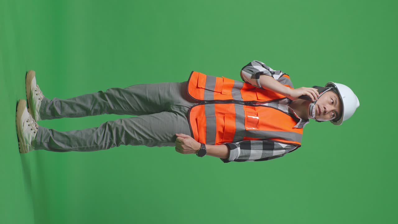 Full Body Side View Of Asian Male Engineer With Safety Helmet Talking On Smartphone While Standing In The Green Screen Background Studio