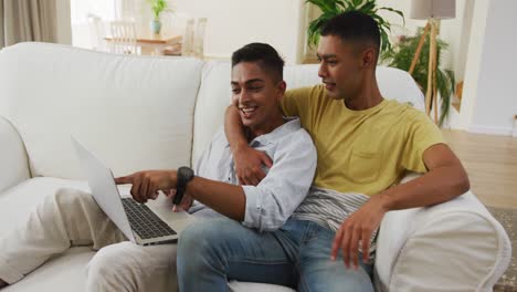 Smiling-mixed-race-gay-male-couple-sitting-on-sofa-looking-at-laptop-and-talking