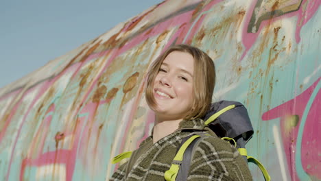 Portrait-Shot-Of-A-Pretty-Girl-With-Backpack-Leaning-On-A-Graffiti-Wall,-Looking-At-The-Camera-And-Smiling