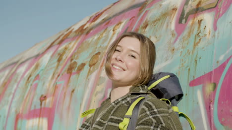 portrait shot of a pretty girl with backpack leaning on a graffiti wall, looking at the camera and smiling