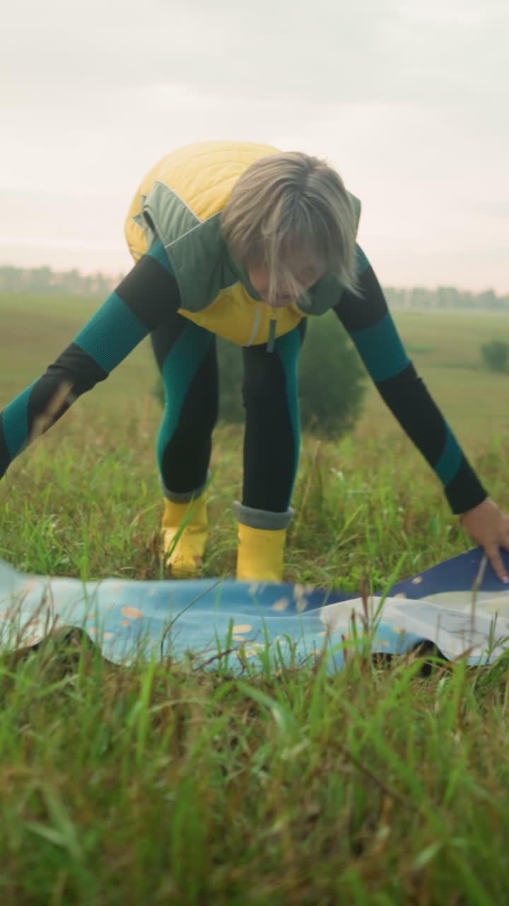 노란색 재과 부츠를 입은 여자가 잔디에 매트를 펴고, 멀리서 나무와 구름이 쌓인 하늘이 있는 광활한 야외에서 야외 활동을 준비하고 있습니다.