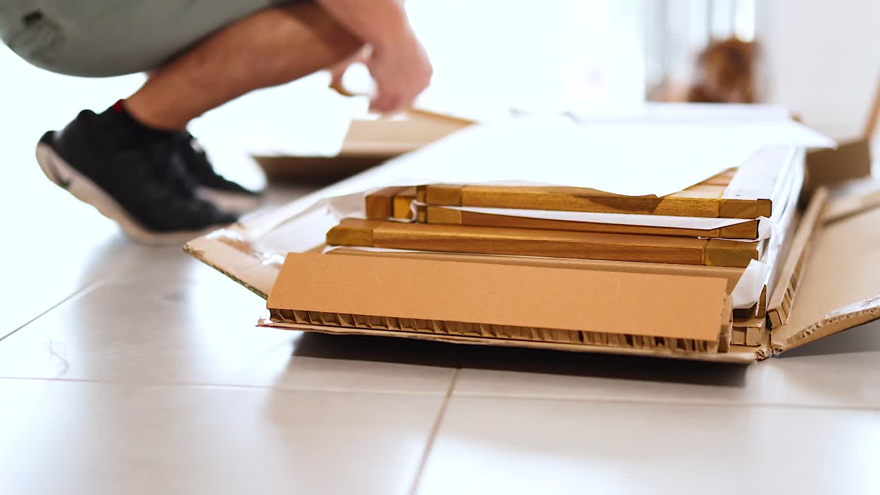 A person unpacks a cardboard box on a tiled floor, revealing contents in a well-lit room