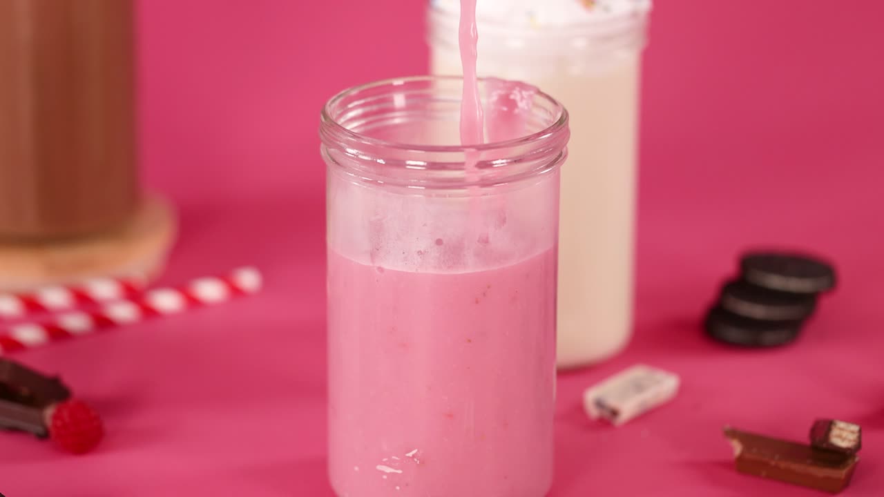 Strawberry milkshake poured into glass jar, surrounded by cookies, candy, and chocolate, pink background