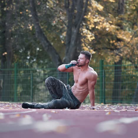 muscular sportsman outdoors. topless man with sportive body having break in the stadium. handsome athlete sits on autumn trees background.