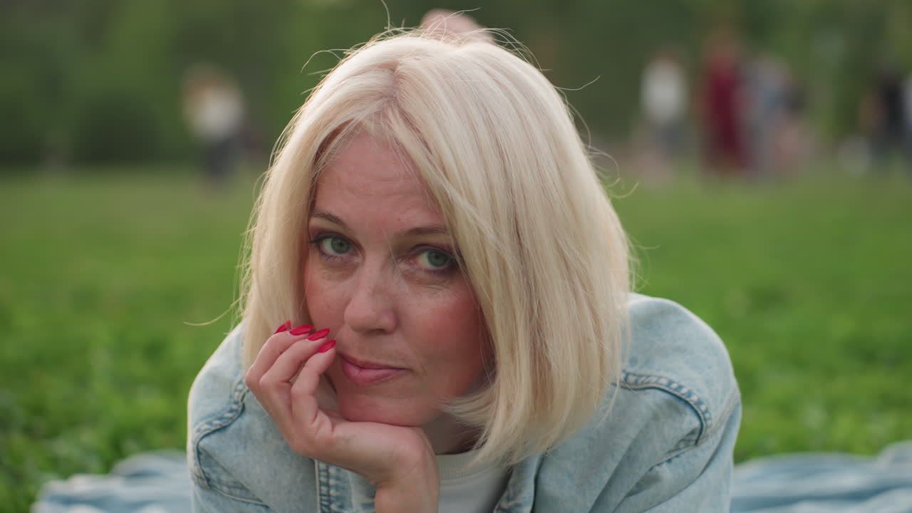 Donna alla moda sdraiata su una coperta nel parco che si sistema i capelli mentre si sostiene la testa con una mano, sorridendo dolcemente, persone sfocate che giocano sullo sfondo, luce calda che crea un'atmosfera calma e rilassata