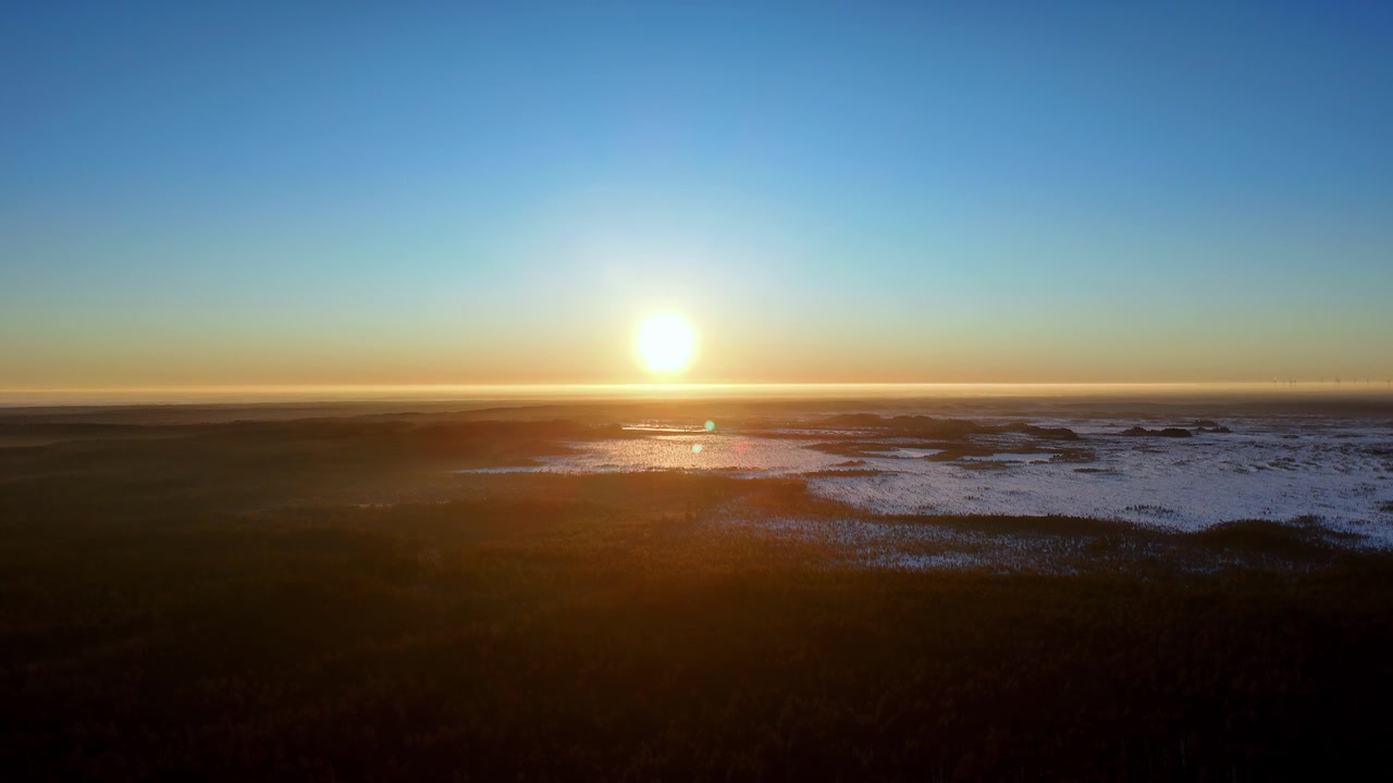 Aerial panoramic vista showcasing a serene winter sunrise over a vast frozen wetland landscape with golden light reflecting on icy patches under a clear blue sky captured from a high angle view
