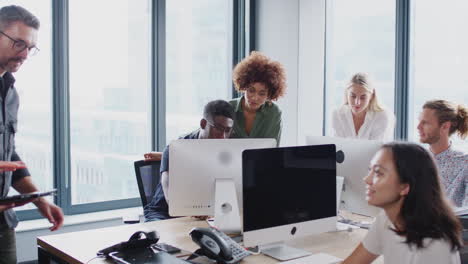 three creatives talking in a busy office, other colleagues working at desks behind them, zoom out