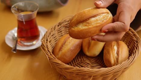 la mano de una mujer se extiende en una canasta de panes, con un vaso de té turco en la mesa