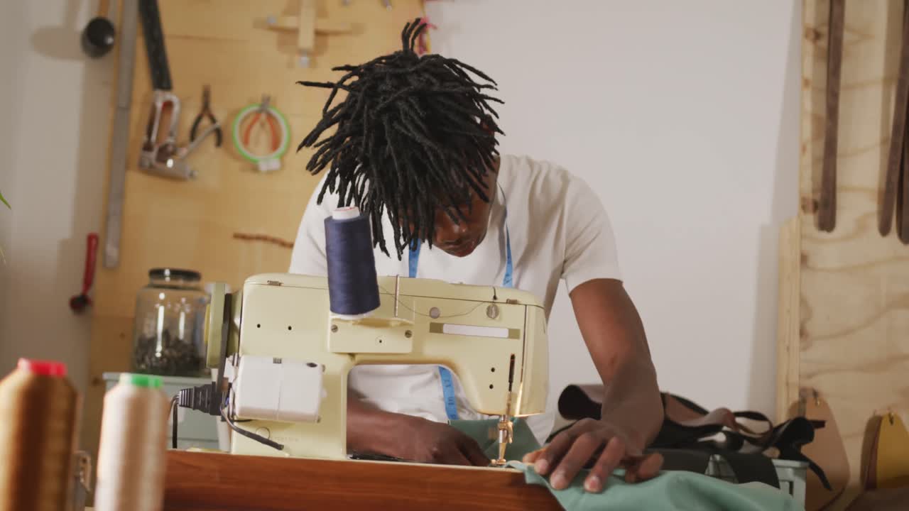 artesano afroamericano enfocado con dreadlocks usando máquina de coser en taller de cuero