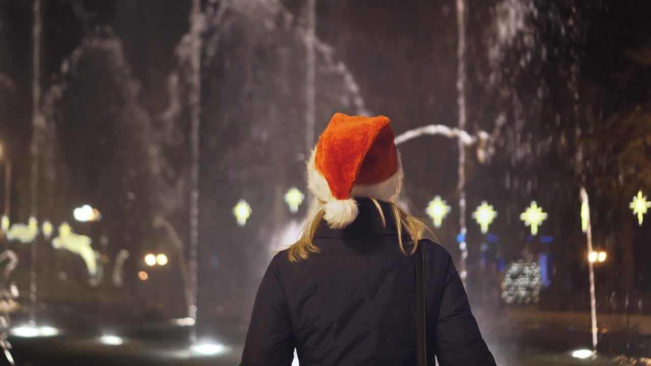 한 소녀 가 산타클로스 모자 를 입고 크리스마스 밤 에 분수 를 바라보고 있다