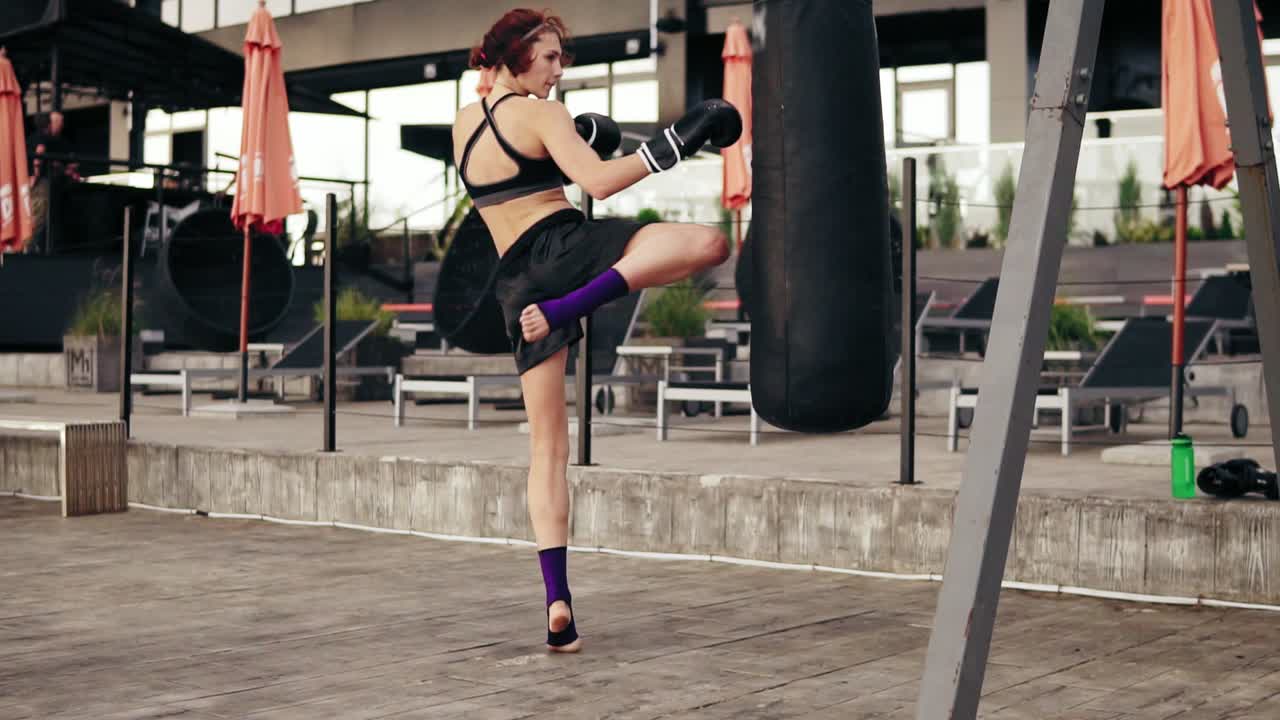 boxeadora atlética fuerte con guantes haciendo ejercicio con una bolsa. golpea una bolsa con la pierna. ejercicio afuera. boxeadora