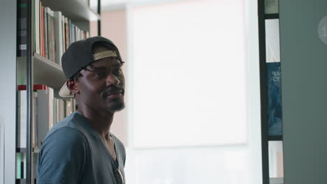 Side view of young man wearing cap backward standing in library aisle, turning with gentle smile as sunlight streams through window, surrounded by tall shelves filled with colorful books