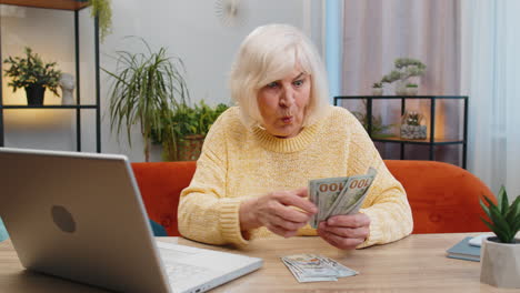 happy senior woman counting money cash and use laptop pc calculate domestic income earnings at home