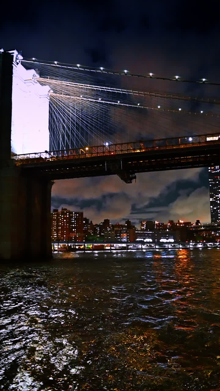 Skyline di New York, la città che non dorme mai, vista sul paesaggio acquatico dell'East River vicino al ponte di Brooklyn. Angolazione dal basso verso il cielo nuvoloso sopra la metropoli notturna. Video verticale