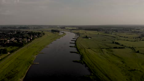 Movimiento-Aéreo-Ascendente-Hacia-Atrás-Que-Muestra-El-Valle-De-Ijssel-Con-Un-Río-Serpenteante-Y-Llanuras-Aluviales-En-Un-Lado-Y-Una-Zona-Residencial-En-El-Otro-Lado-En-Los-Países-Bajos-En-Un-Día-Nublado-Durante-La-Puesta-De-Sol