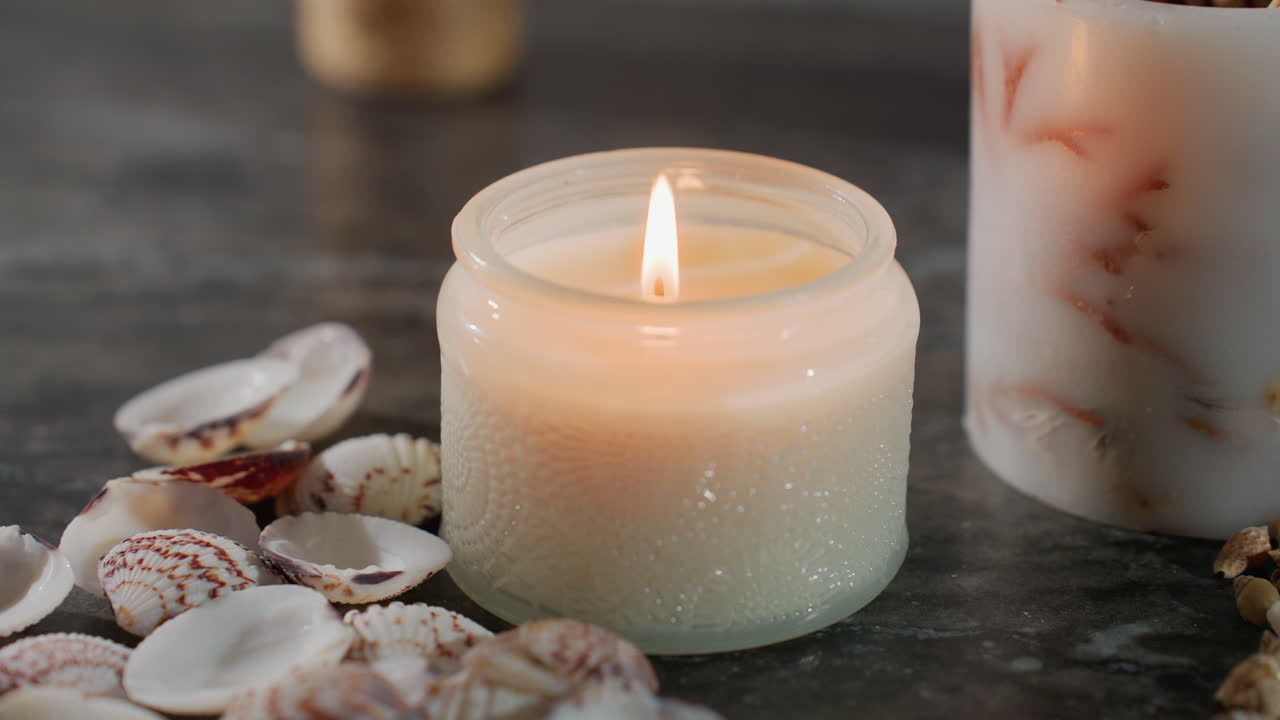 White candle burning steadily in glass jar surrounded by seashells and starfish on dark marble surface, soft glow illuminating elegant cozy setting with natural elements