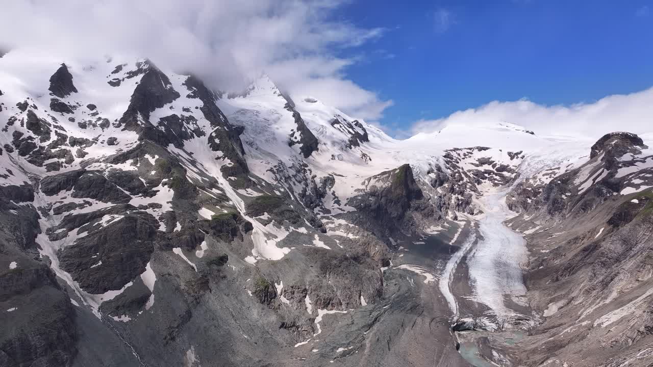 Aerial panorama of Großglockner high alpine region surrounded by rugged mountains and blue sky, white snow accents grey rocks with icy valley, Austria landscape view