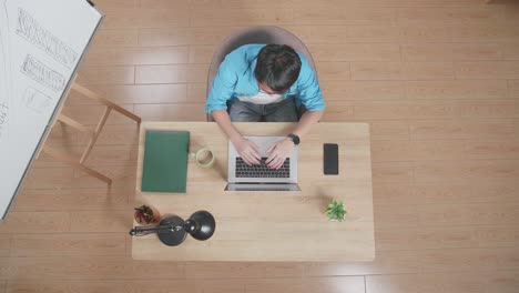 top view of a man stretching while sitting at the wooden desk besides a whiteboard typing on a laptop next to the smartphone at home