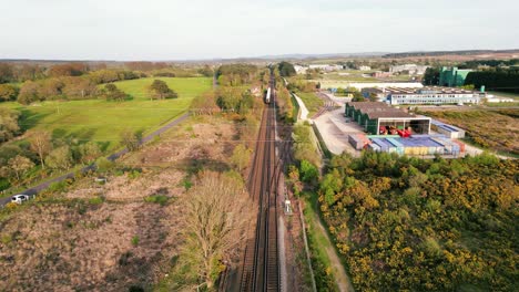 Tren-Retrocediendo-En-La-Distancia-En-Una-Vía-Con-El-Campo-A-Un-Lado-Y-El-Almacén-Y-El-Polígono-Industrial-Al-Otro.