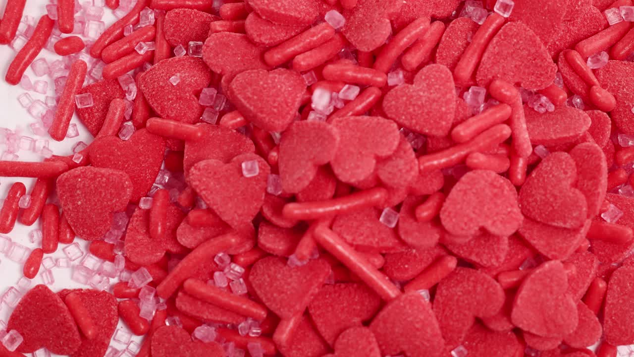 Red heart-shaped candy sprinkles cascade onto a surface, creating a vibrant, textured pile in bright lighting