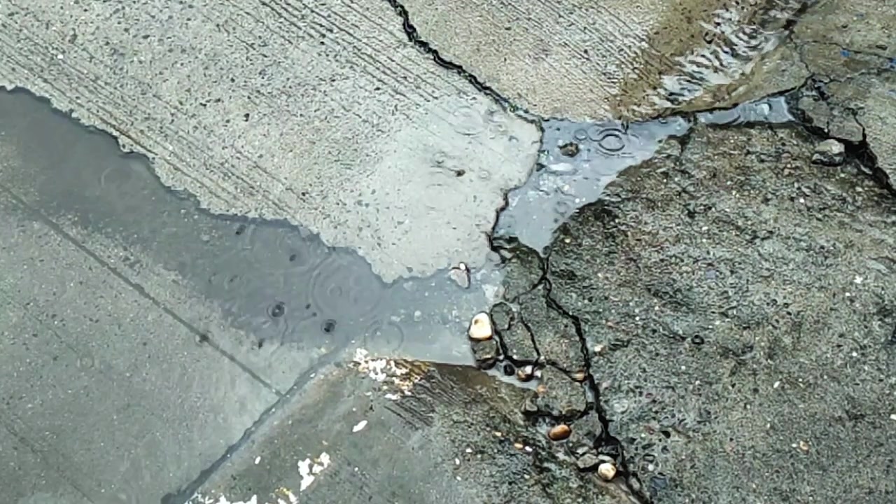 Gocce di pioggia che colpiscono il marciapiede bagnato al rallentatore, creando increspature e piccoli schizzi su una superficie di cemento fessurata durante il tempo piovoso.