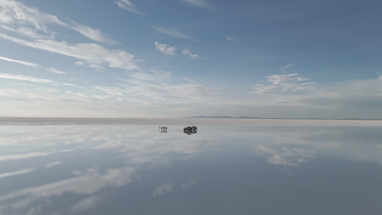 Drone shot flying above the mirror salt flats in Bolivia Uyuni during sunset with the visible reflection and a car with tourists around it LOG